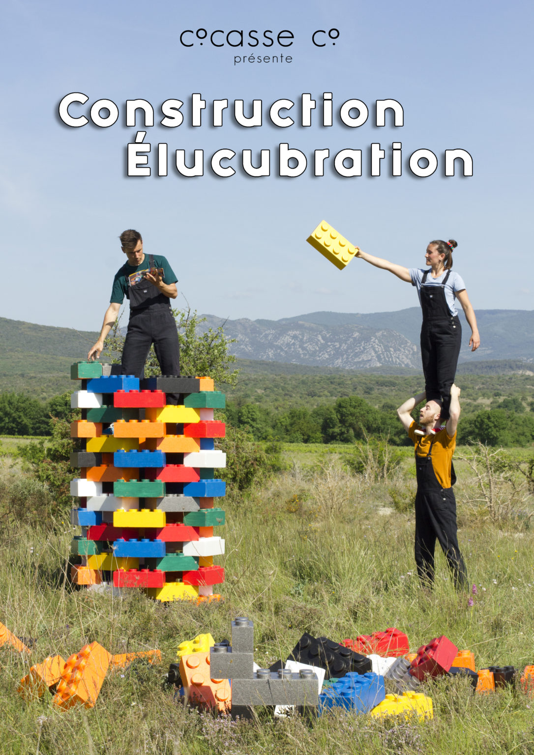 Construction Elucubration / Cie Cocasse / Une personne en porte une sconde sur ses épaule debout, elle tient un immense lego qu'elle tend à une personne en hauteur sur une immense tour de lego. Le tout dans un grand champ